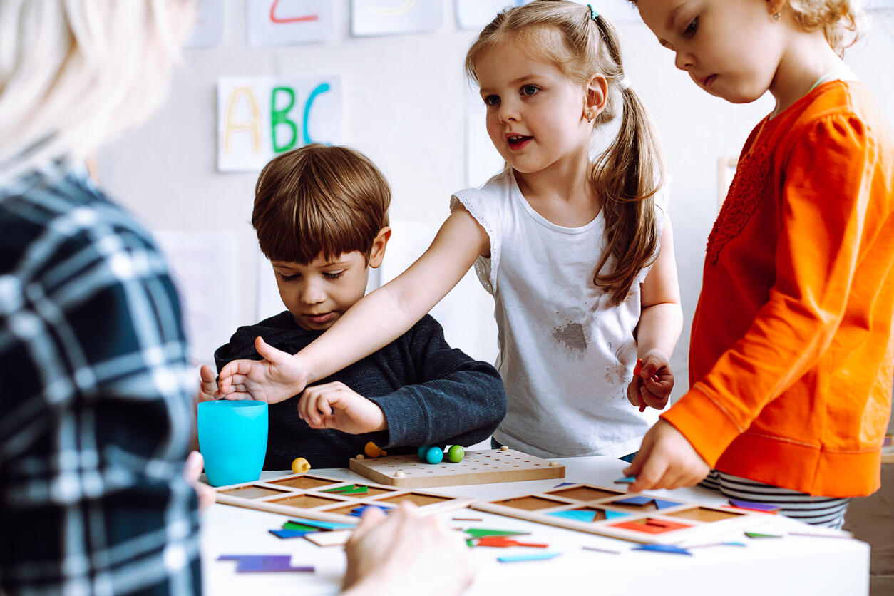 Two beautiful little girls standing at table with pencils in bright classroom. Little boy sitting, playing board games.